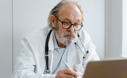 Elderly doctor with glasses and stethoscope working on a laptop in a medical office.